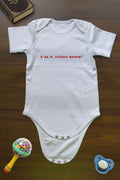 White baby onesie with 'I'm a Jesus baby!' text on a wooden surface with toys.